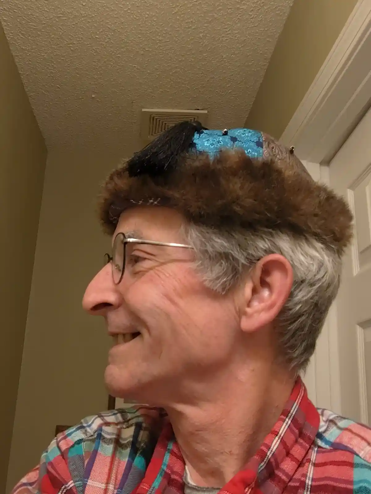 A smiling older man with glasses and gray hair wears a brown fur-trimmed hat with a blue patterned top and black tassel. He…