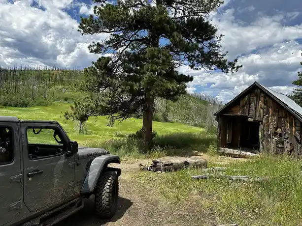 A gray, mud-splattered Jeep Wrangler Rubicon is parked on a dirt path next to a dilapidated wooden shed in a grassy field…