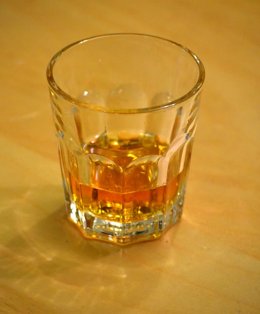 A close-up photo shows a faceted, clear glass tumbler containing a small amount of amber-colored liquid, likely whiskey…
