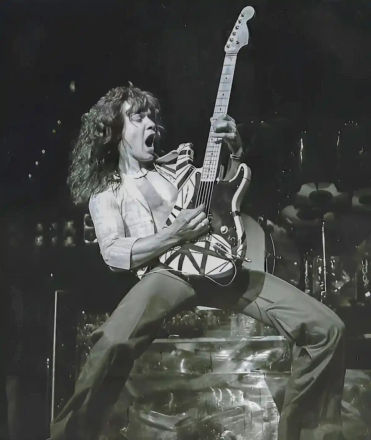 A black-and-white photo shows a male musician with long curly hair, wearing a partially unbuttoned shirt and pants, sitting…