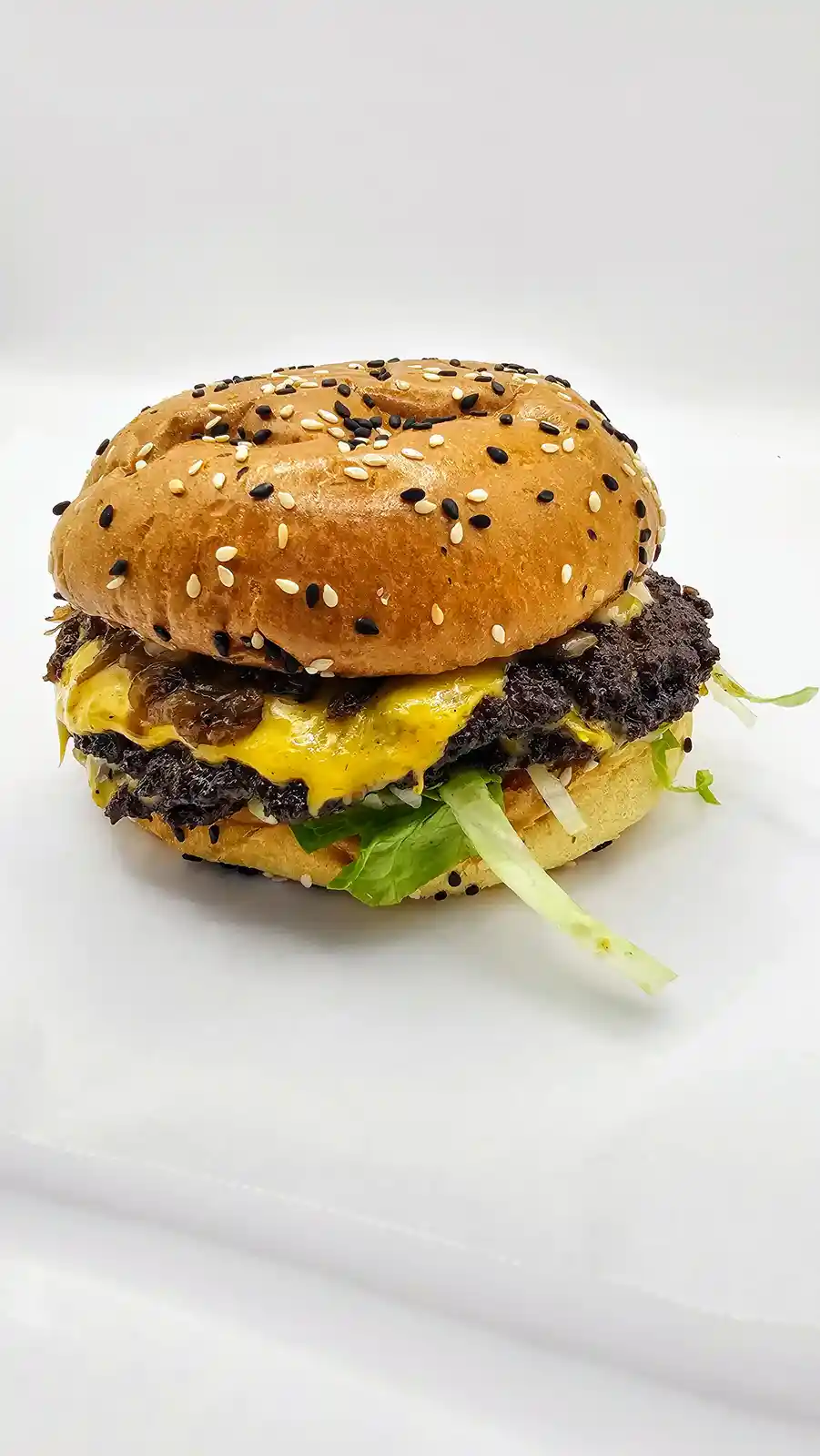 A close-up photo of a cheeseburger with a sesame seed bun, featuring two dark, charred-looking beef patties, melted yellow…