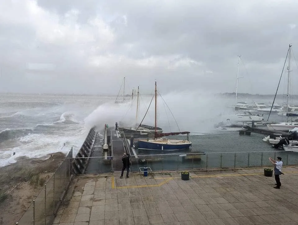 A stormy harbor scene with large waves crashing against a pier, sending spray into the air. Several sailboats are docked…
