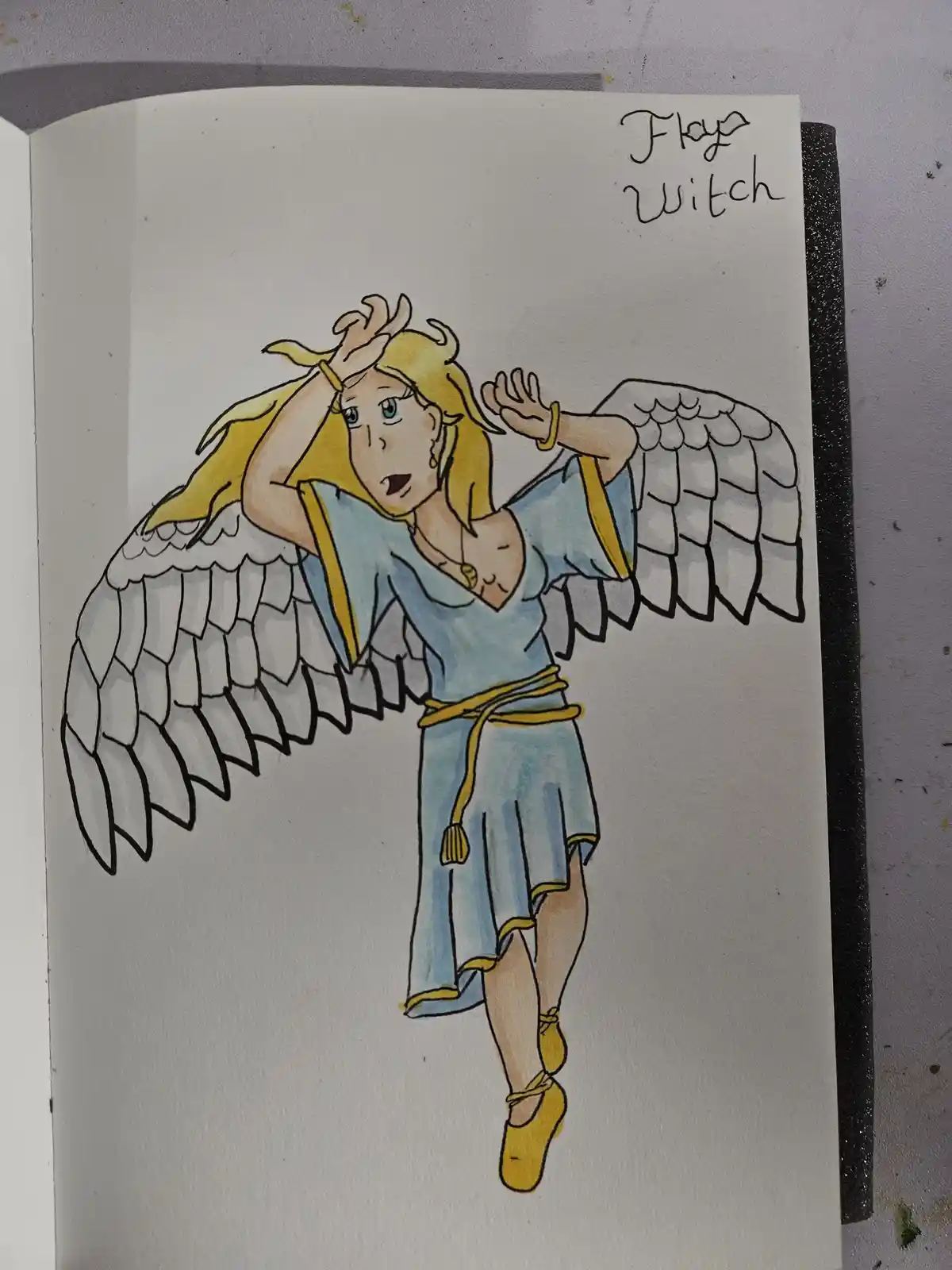 A hand-drawn illustration in a sketchbook shows a blonde-haired female character with large, detailed white angel wings…