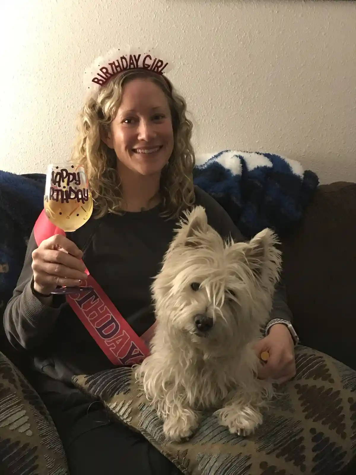 A smiling woman with curly blonde hair sits on a couch wearing a 'BIRTHDAY GIRL' headband and a red 'BIRTHDAY' sash, holding…