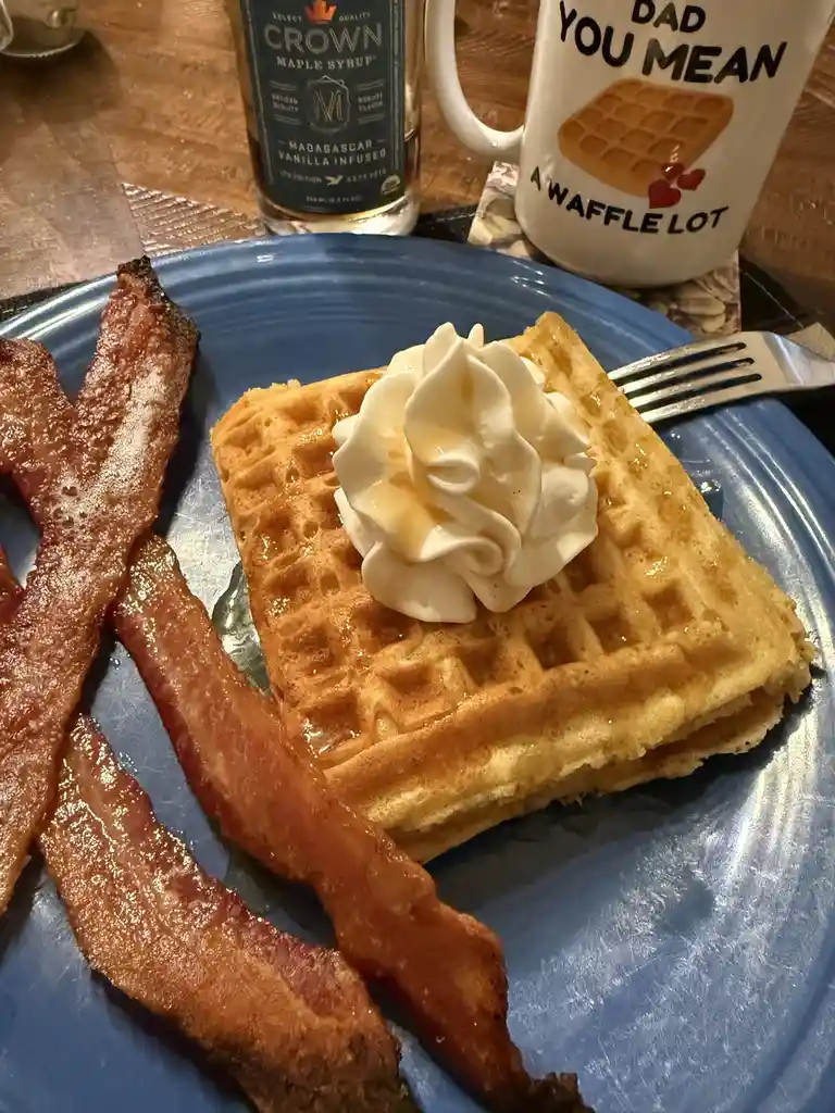 A close-up photo of a breakfast plate with a golden-brown waffle topped with whipped cream and syrup, accompanied by crispy…