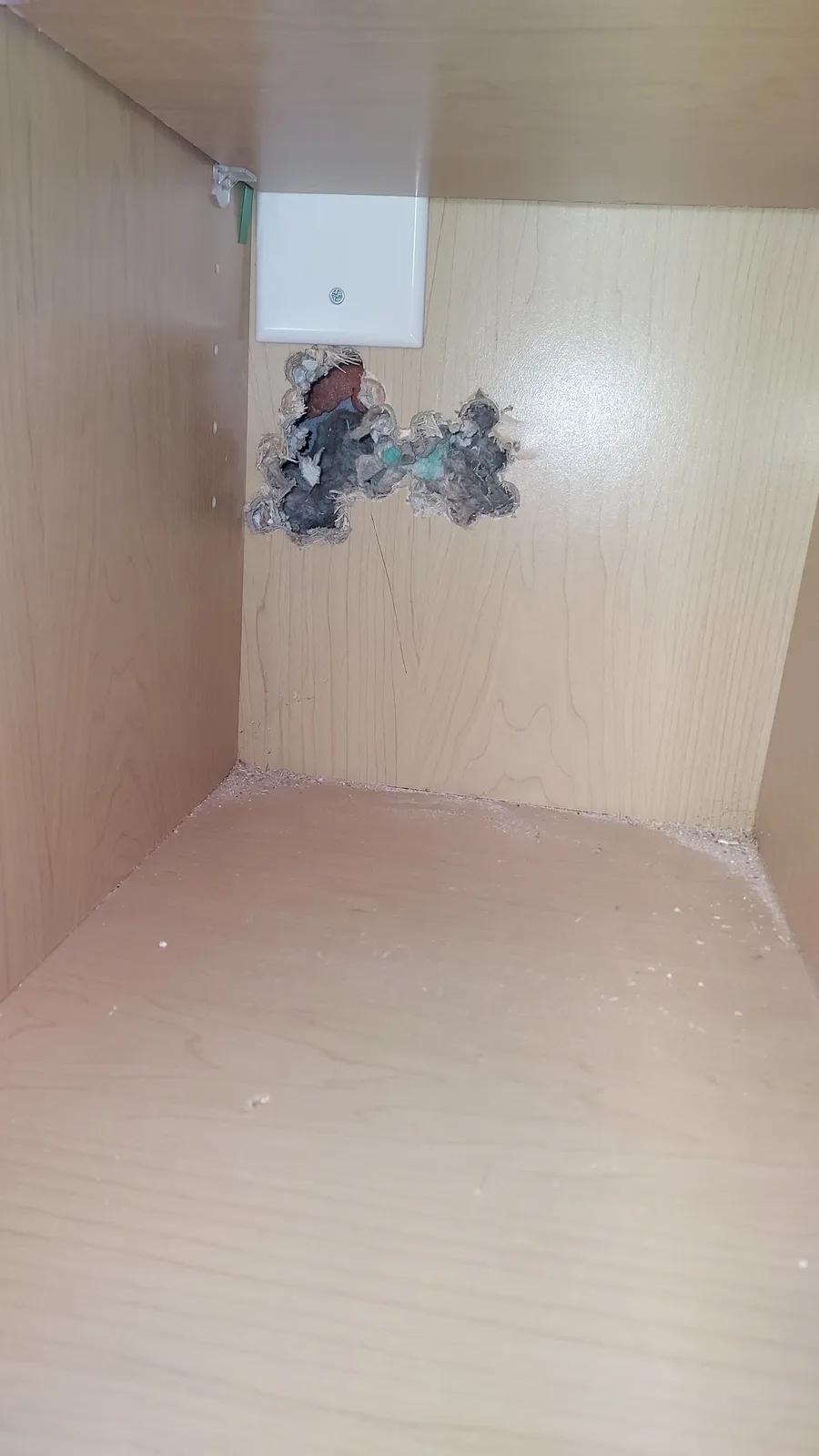 A close-up photo shows the inside of a light-colored wooden cabinet with a large, ragged hole punched through the back…