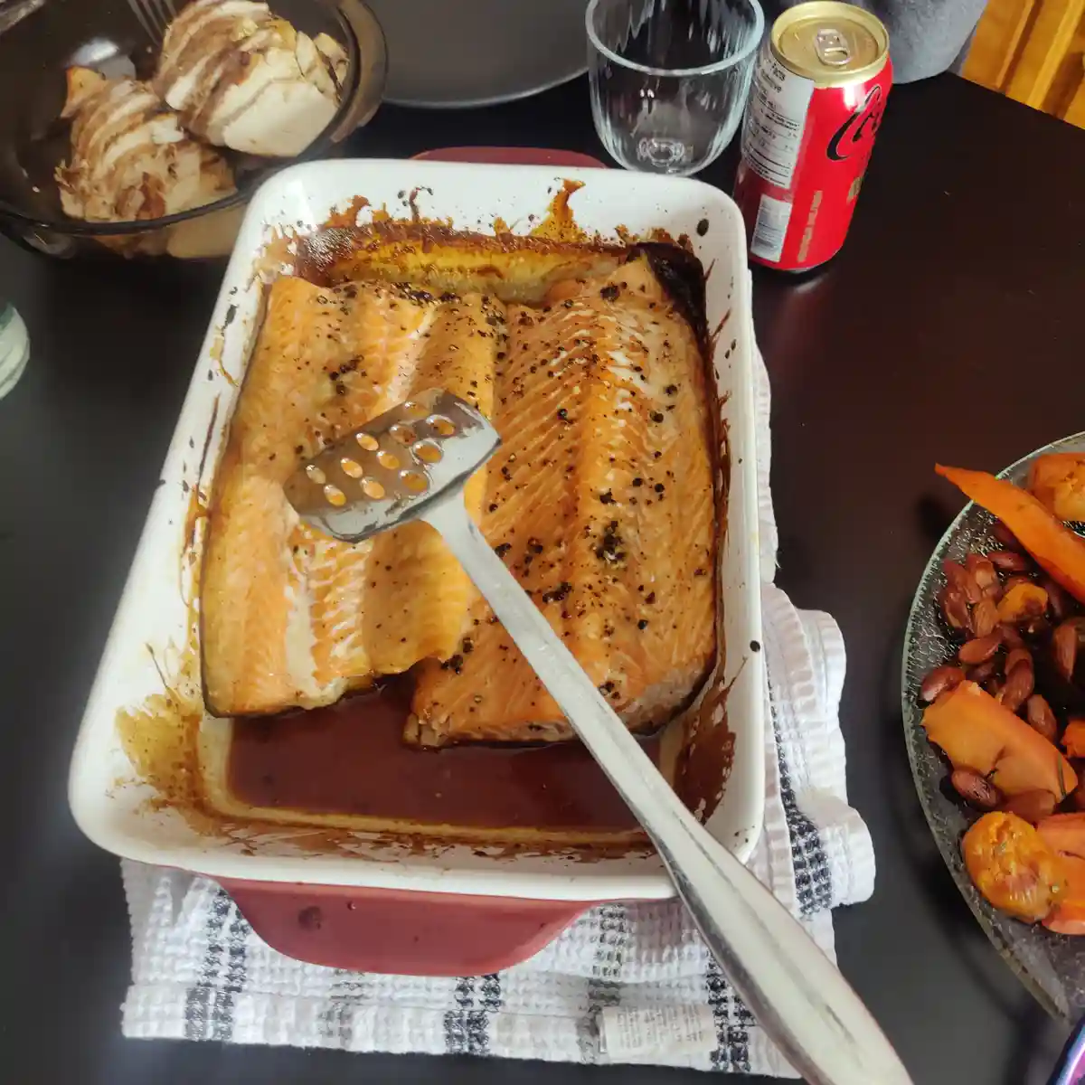 A white baking dish with two cooked salmon fillets sits on a dark table, with a metal spatula resting on top. Surrounding…
