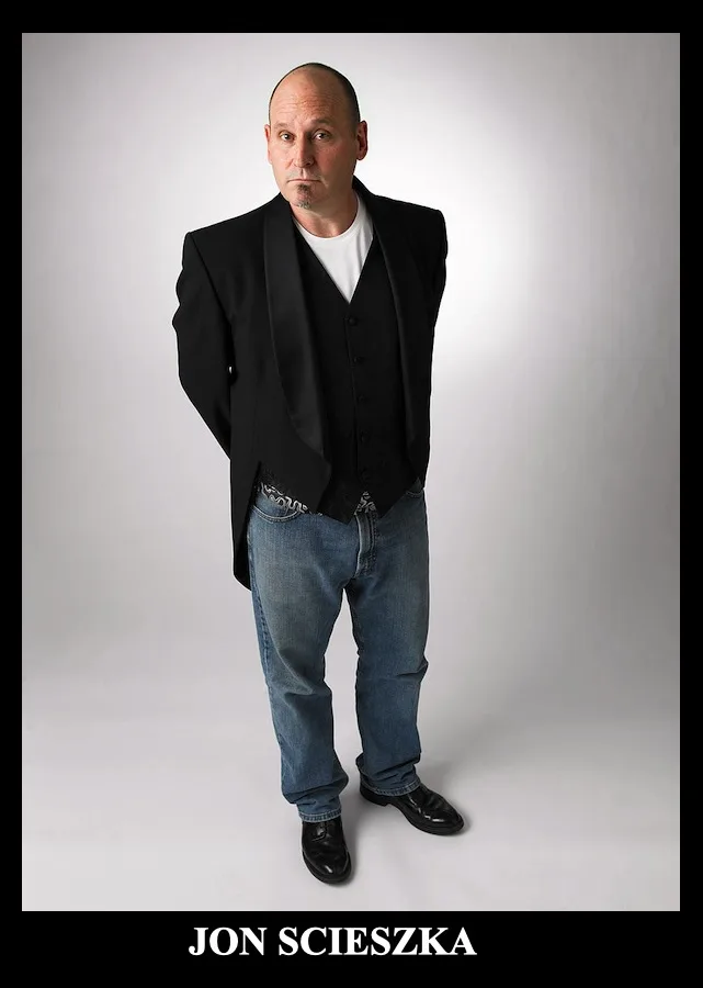 A full-body studio portrait of Jon Scieszka, a bald man with a goatee, wearing a black tuxedo jacket and vest over a white…