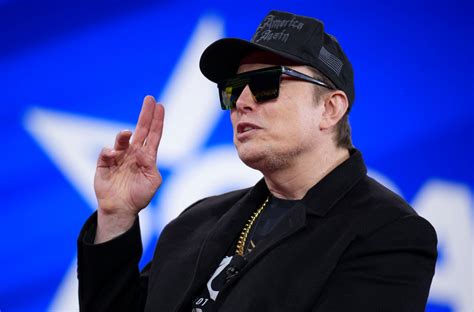 elton john wearing a black cap and sunglasses, making a peace sign with his right hand, standing in front of a blue…
