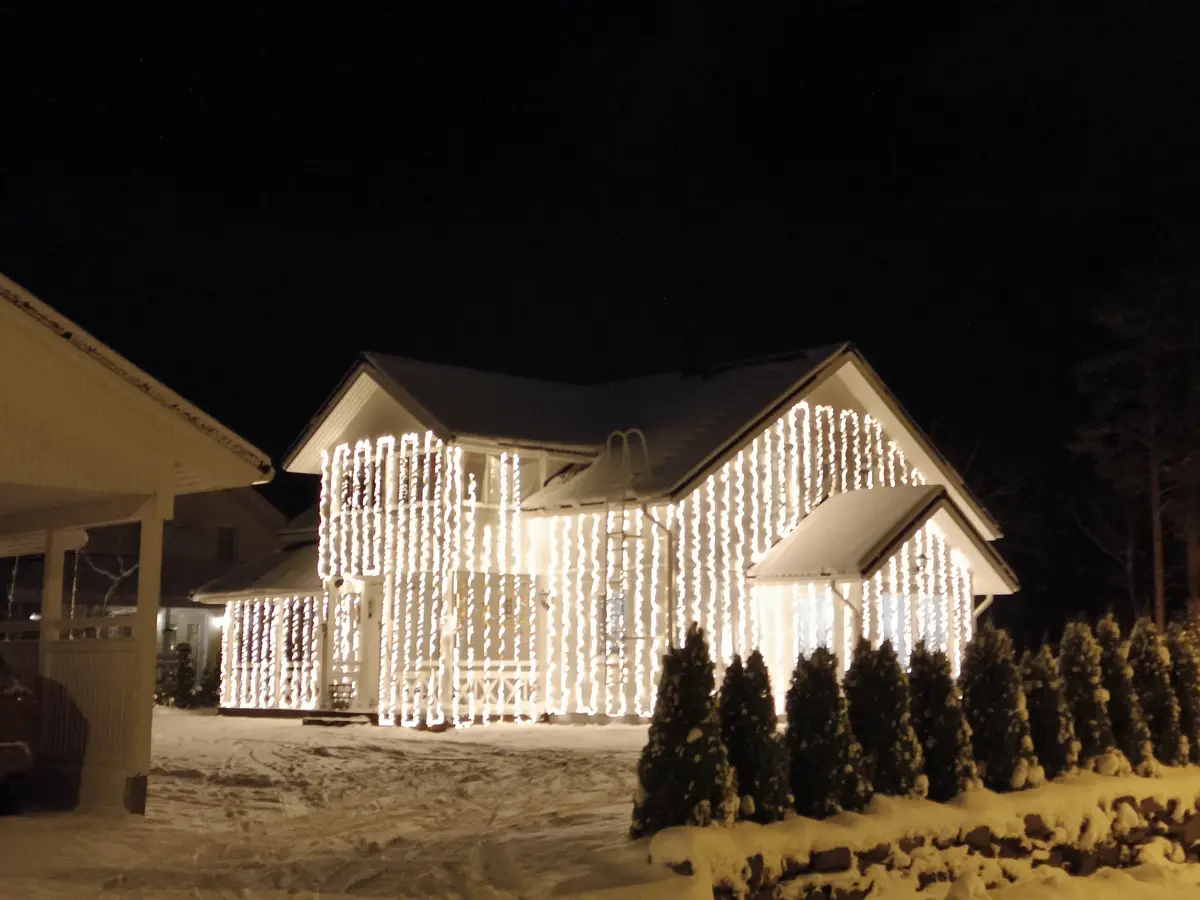 A house covered in vertical strings of bright white Christmas lights stands out against a snowy night, with snow on the roof…