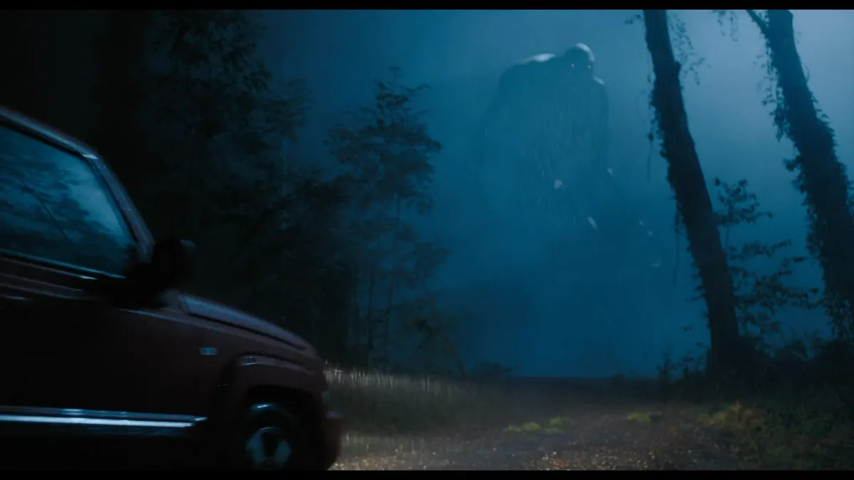 A dark, rainy forest scene shows the front half of a red SUV parked on a dirt road, with a large, shadowy, humanoid monster…
