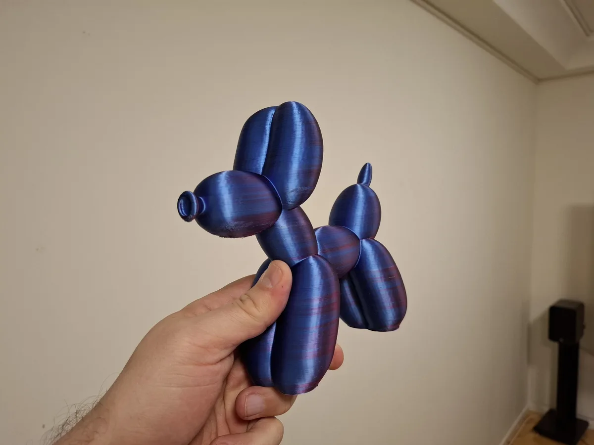 A hand holds a shiny, metallic blue and purple 3D-printed figurine shaped like a balloon animal dog, with visible layer…
