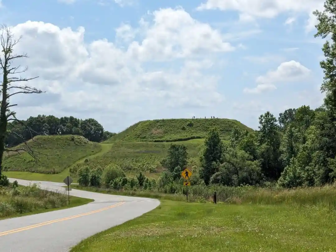 A winding road curves toward large, grassy mounds resembling burial mounds or ancient earthworks, with a yellow pedestrian…