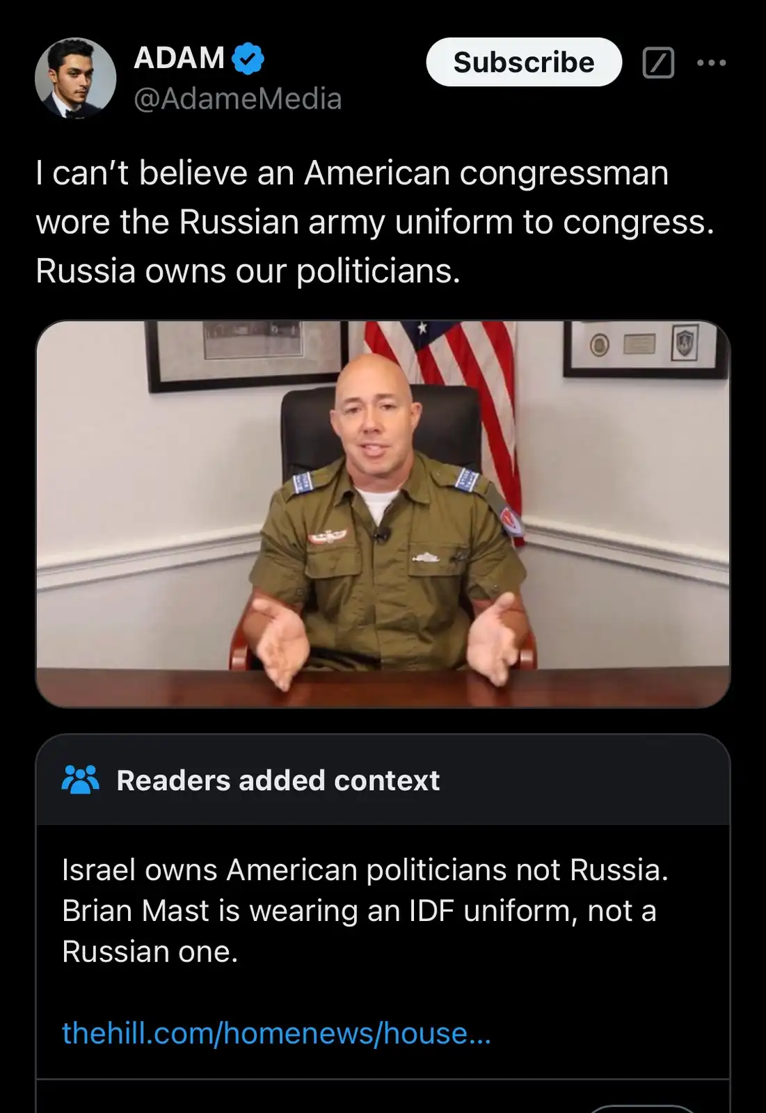 A bald man in an olive green military-style uniform sits at a desk with an American flag behind him. The overlaid text…
