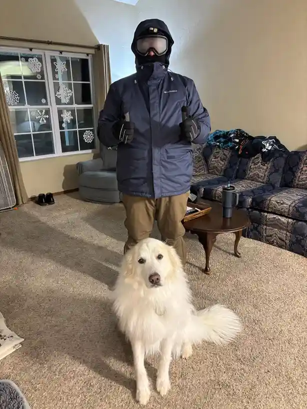 A person wearing full winter gear including a dark blue Columbia ski jacket, goggles, and gloves, stands indoors in a living…