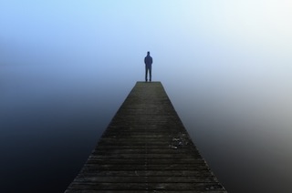 A solitary silhouette of a person stands at the far end of a long, weathered wooden pier extending into a misty, calm body…