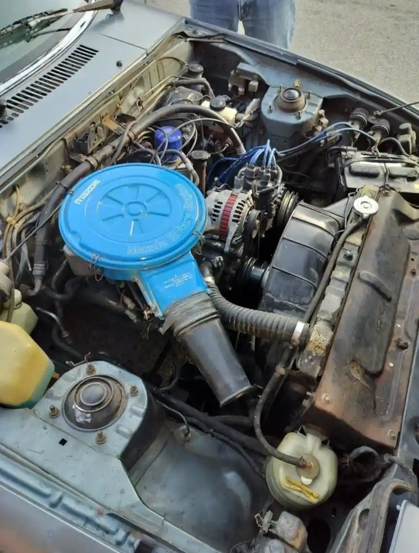 A close-up photo of a car's engine bay, prominently featuring a bright blue Mazda rotary engine cover with 'Mazda Rotary…