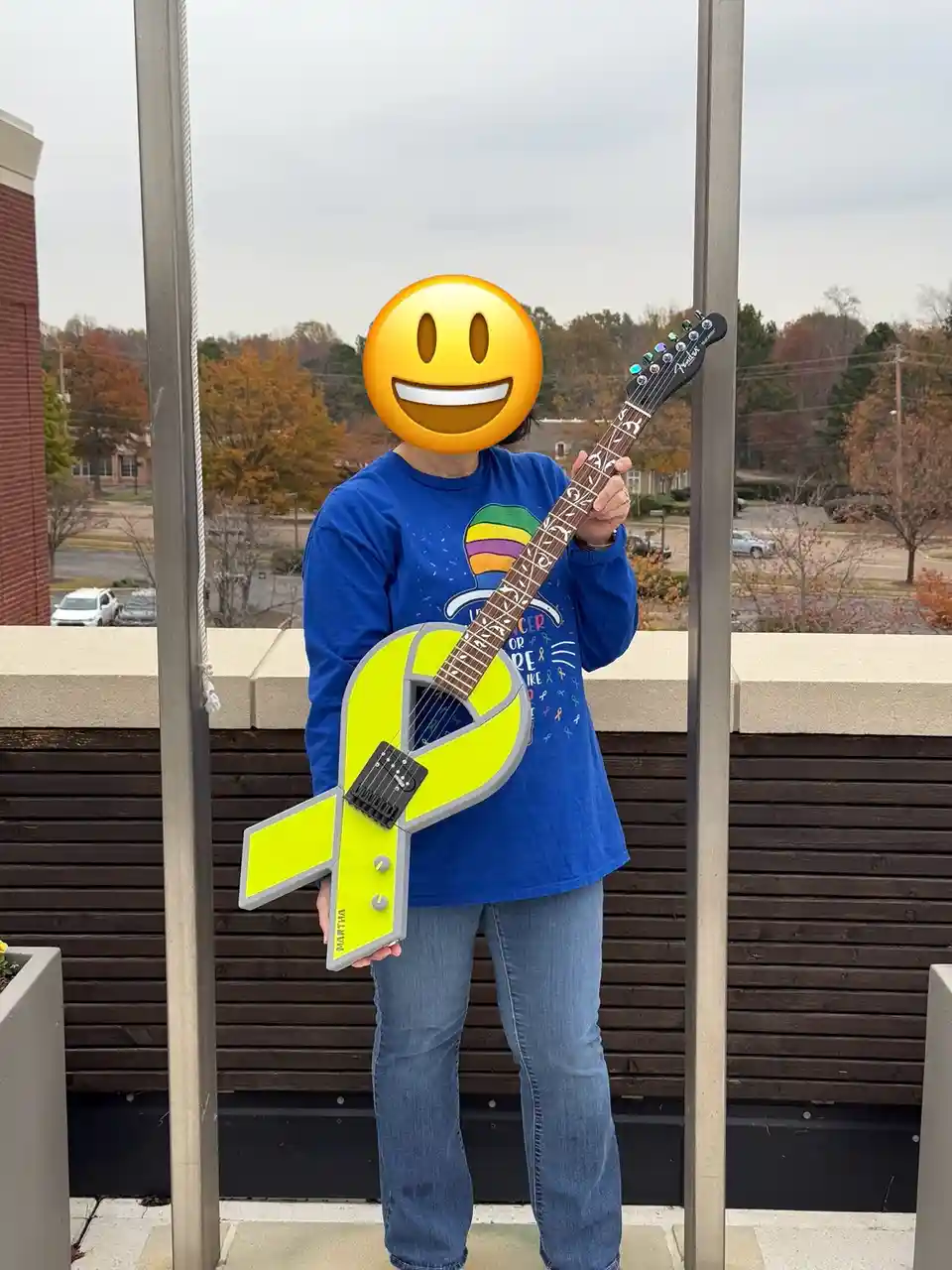 A person with a smiley face emoji covering their face stands on a balcony holding a bright yellow guitar shaped like a…