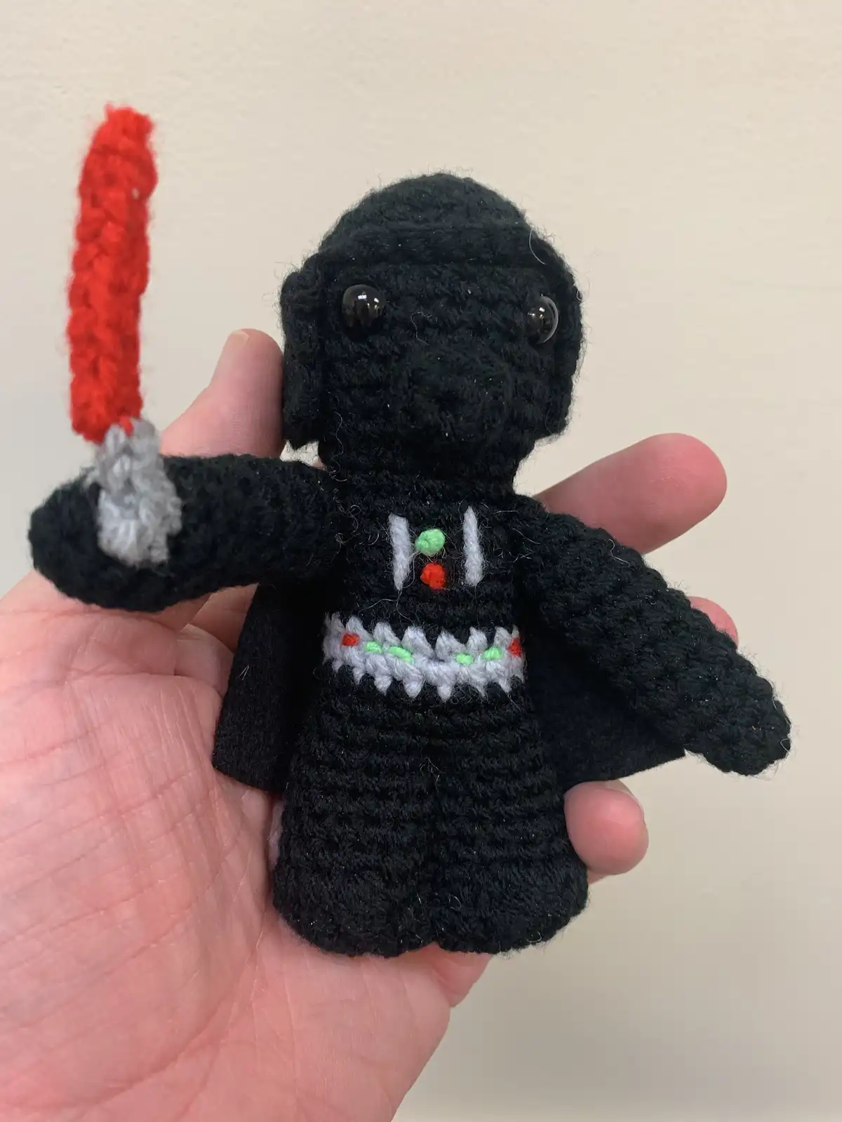 A hand holds a small, black crocheted plush toy resembling Darth Vader, complete with a red crocheted lightsaber. The toy…