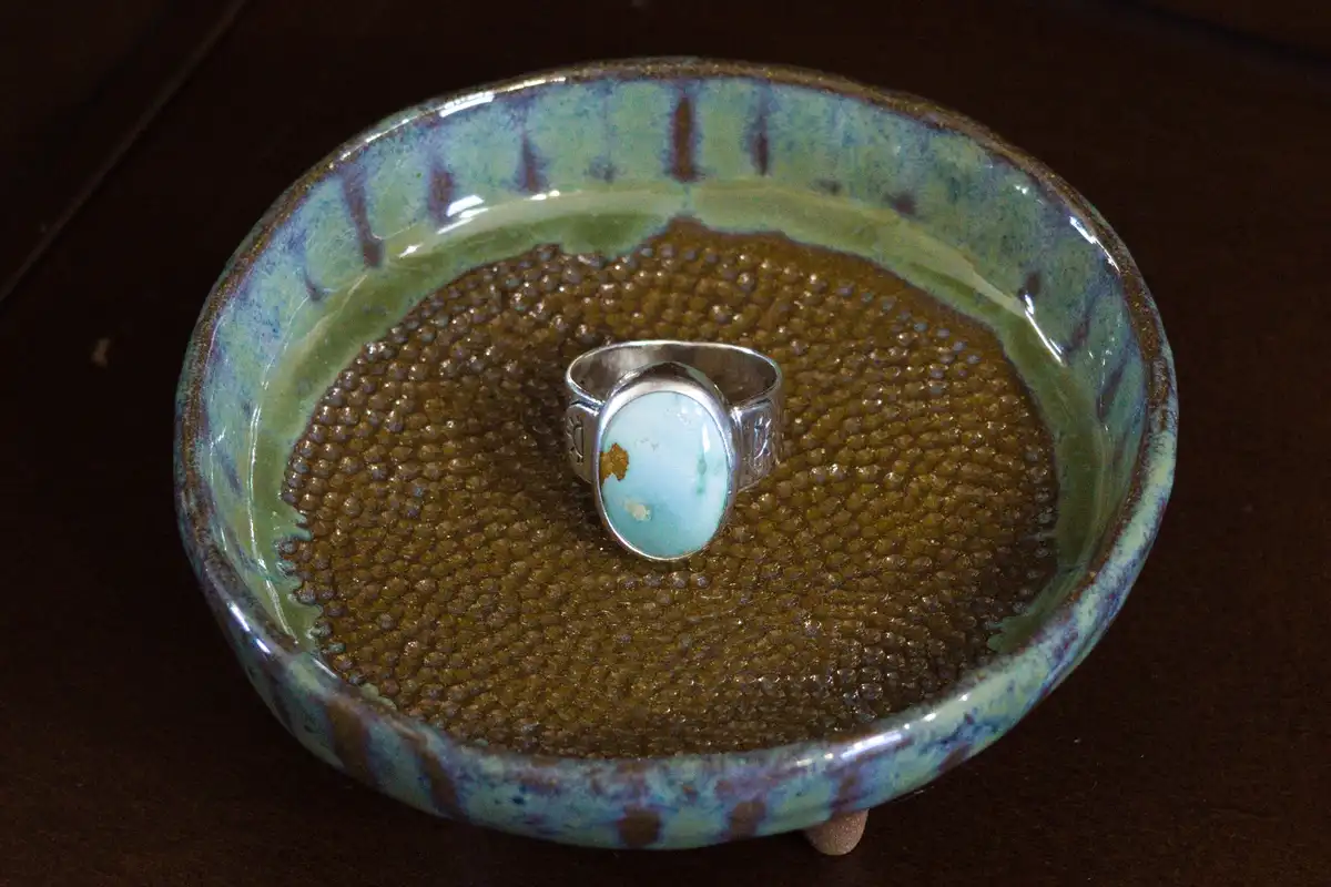 A silver ring with a turquoise stone sits centered in a shallow ceramic bowl filled with small, brown, bead-like grains…