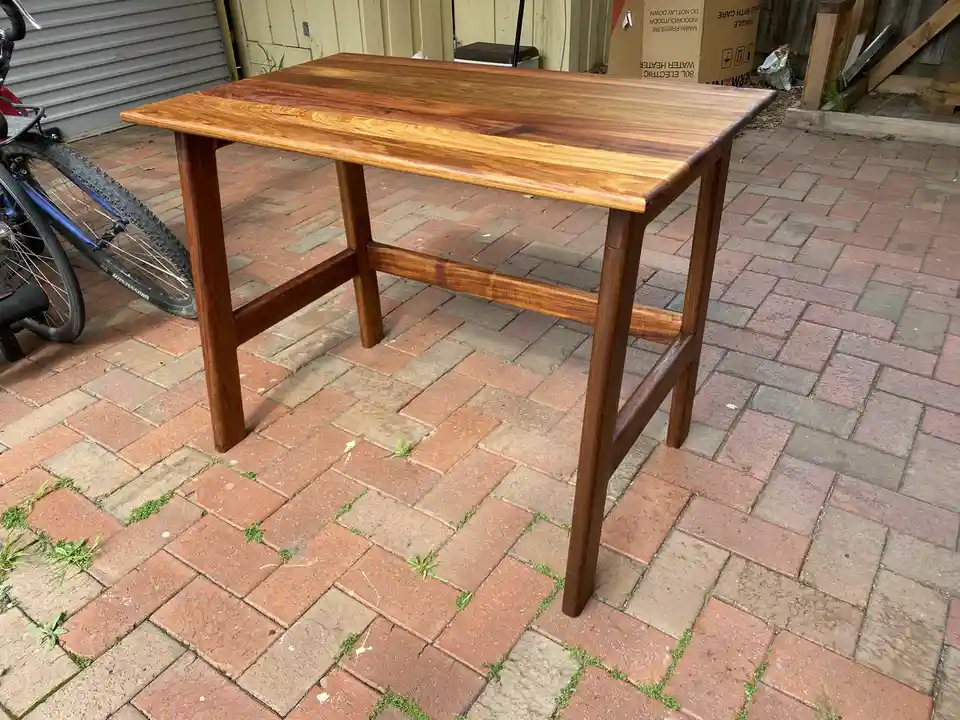 A polished wooden table with four legs and a central stretcher stands on a brick patio where grass grows between the bricks…