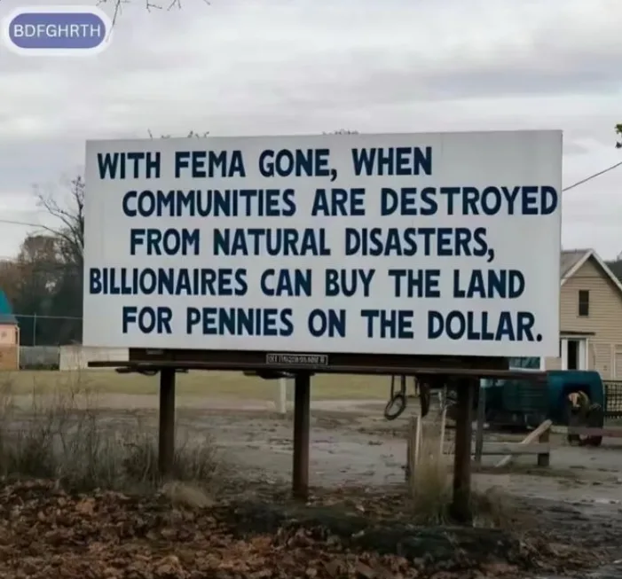 A large white billboard with bold blue text stands in a rural, overcast setting with dry grass and a house in the…