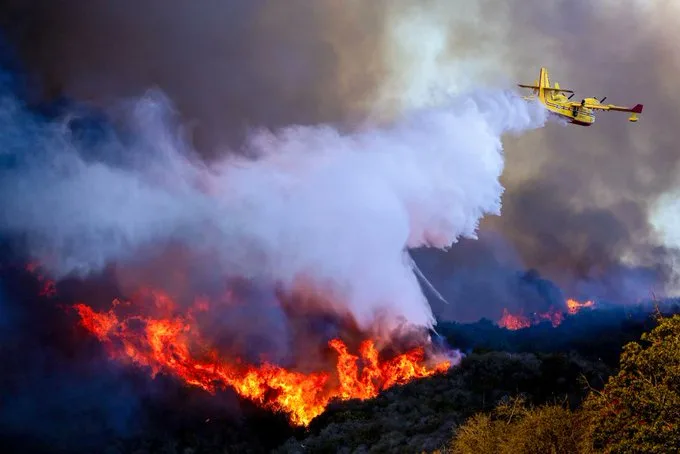 A yellow firefighting aircraft is seen flying low over a raging wildfire, releasing a massive stream of water or fire…
