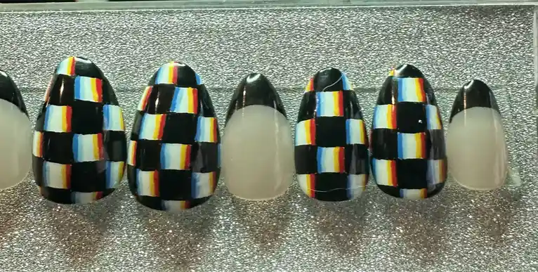 A row of artificial fingernails is displayed on a glittery silver surface. Most nails feature a colorful, checkered pattern…