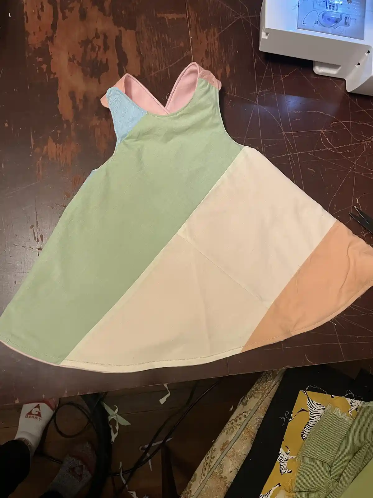 A handmade dress with a patchwork design of pastel green, cream, peach, and light blue fabric lies flat on a scratched…