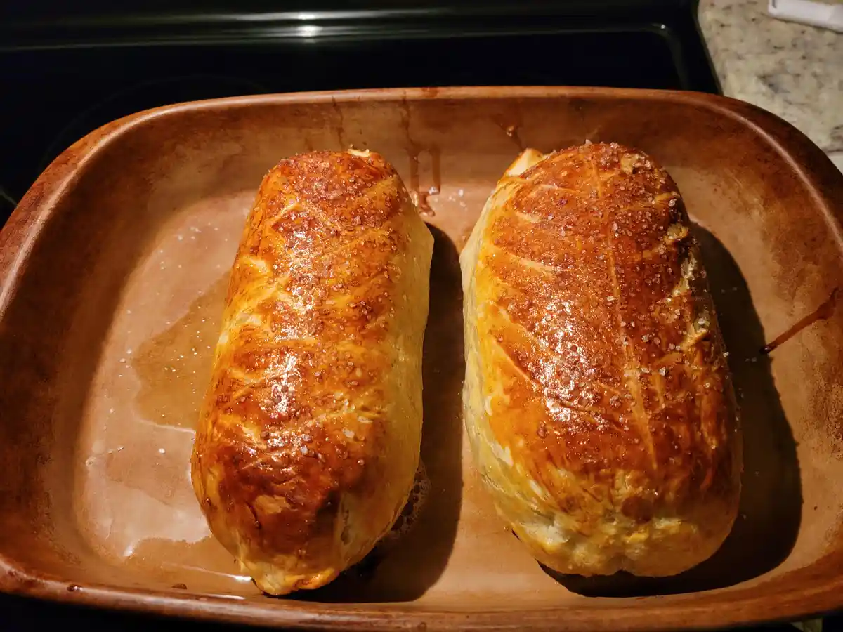 Two golden-brown, flaky pastry rolls sit side-by-side in a brown ceramic baking dish, sprinkled with coarse salt and…
