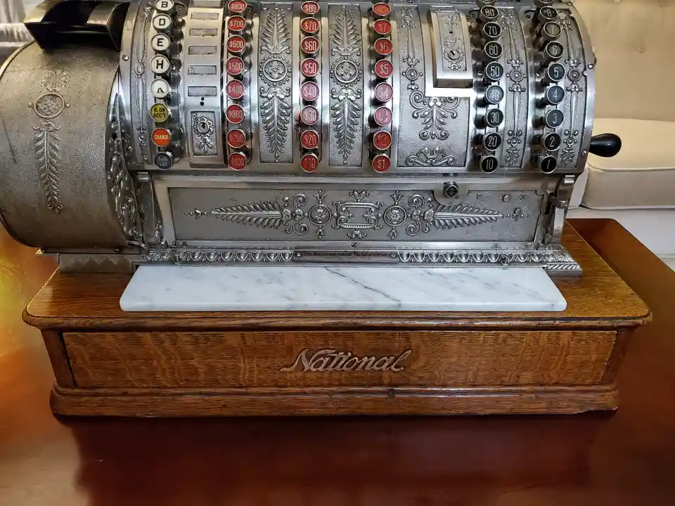 A close-up photograph of an ornate, vintage National Cash Register machine with intricate metalwork, a marble countertop…