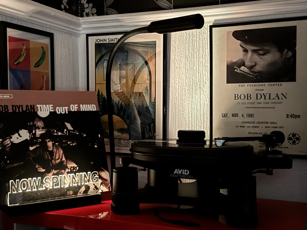 A black turntable sits on a red surface with a glowing sign reading 'NOW SPINNING' in front of it. Behind the turntable are…