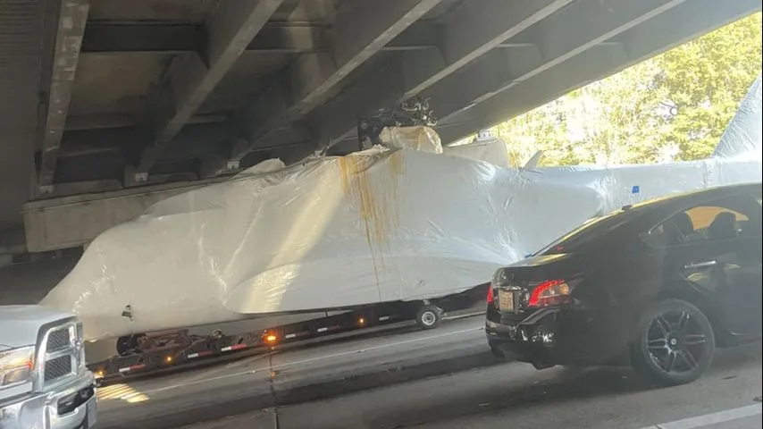 A large white boat covered in plastic is being transported on a trailer under an overpass, with a black sedan partially…