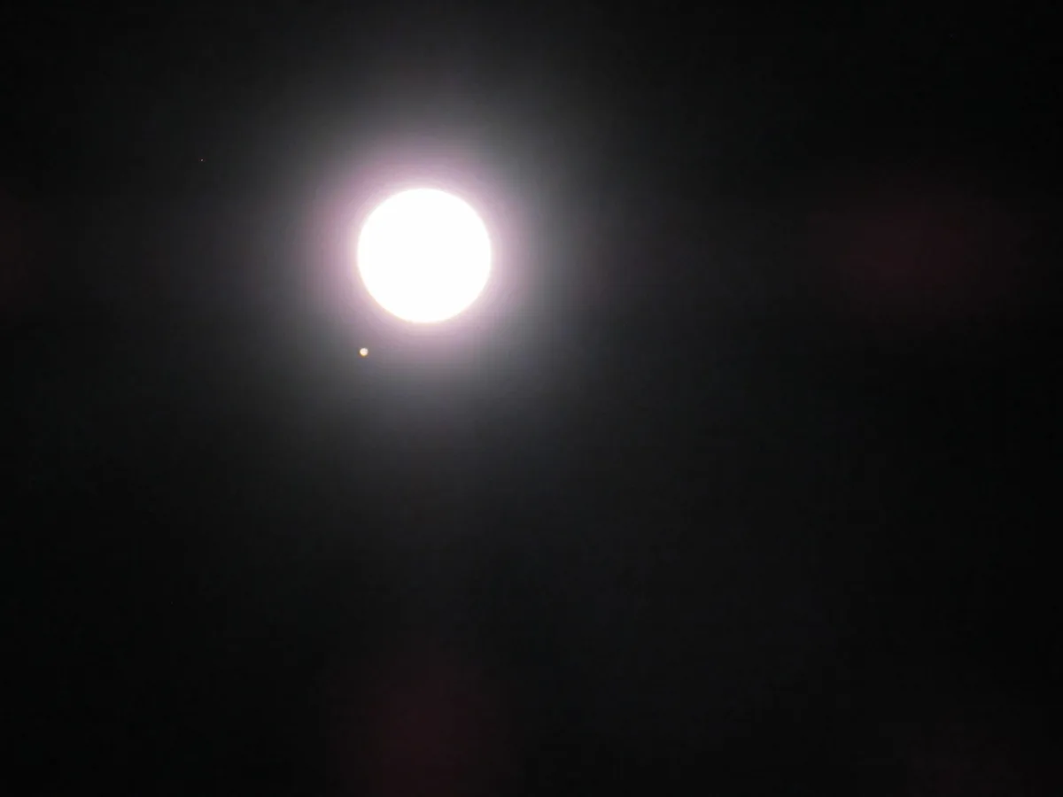 A bright, overexposed full moon dominates a dark night sky, with a small, dim star or planet visible nearby. The image has…