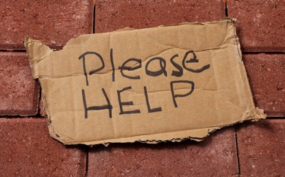 A torn piece of brown cardboard with handwritten text 'Please HELP' in black marker lies on a red brick pavement. The text…