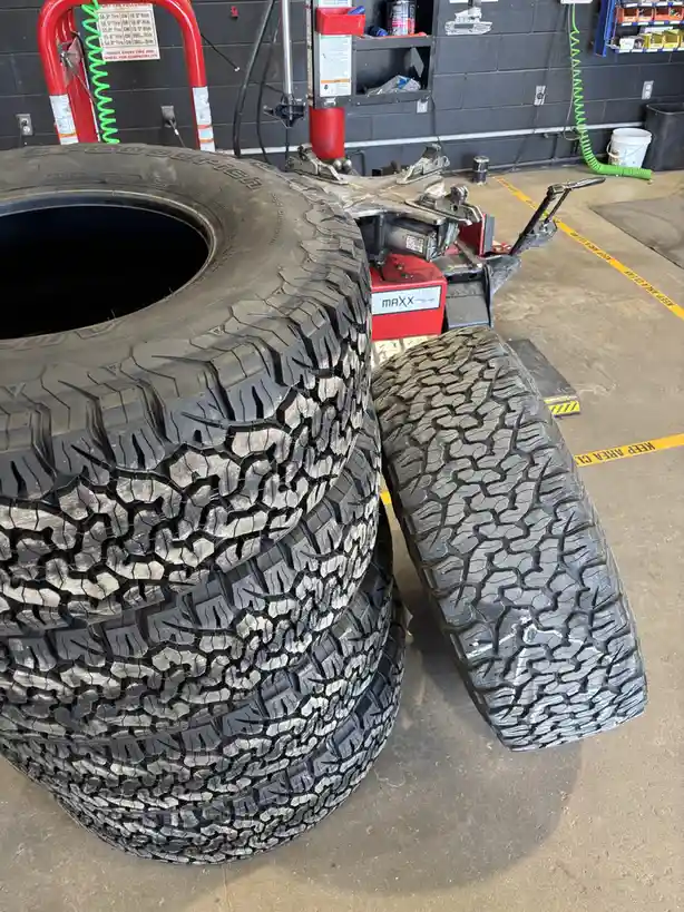 A photo shows four large, rugged off-road tires stacked in a garage, with one tire lying beside the stack. The background…