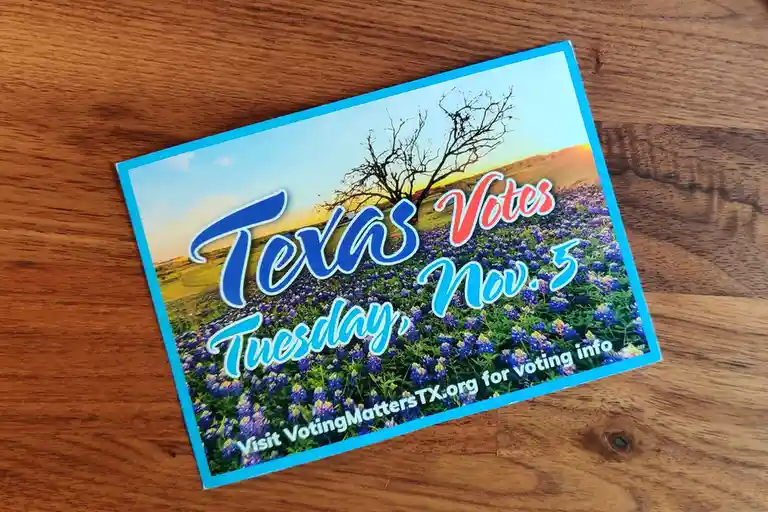 A rectangular political sticker lies on a wooden surface, featuring a scenic image of a field of bluebonnets under a sunset…
