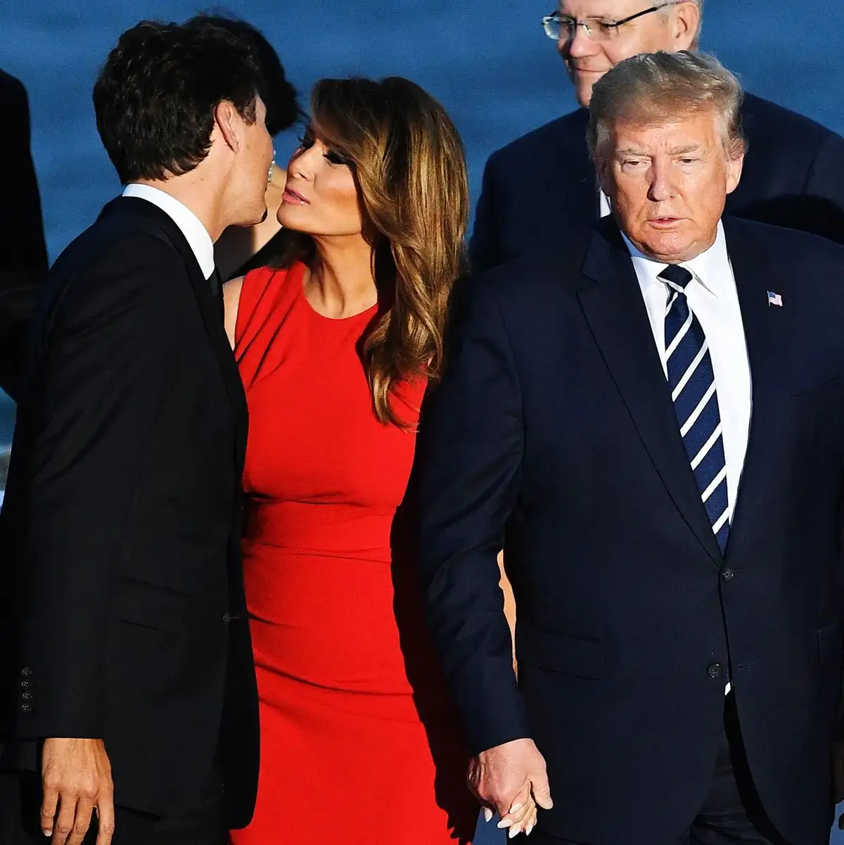 A photo shows Melania Trump in a red dress whispering to a man in a black suit while holding hands with Donald Trump, who…