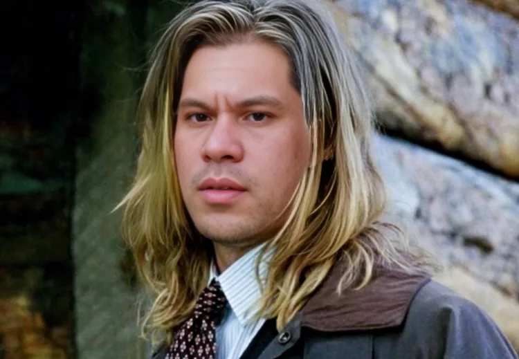 a man with long blonde hair, wearing a collared shirt and patterned tie under a jacket, stares intensely at the camera with…