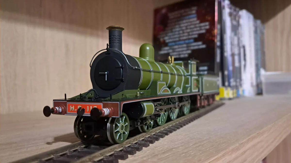 A close-up photograph of a detailed green and black model steam locomotive on a miniature track, resting on a wooden shelf…