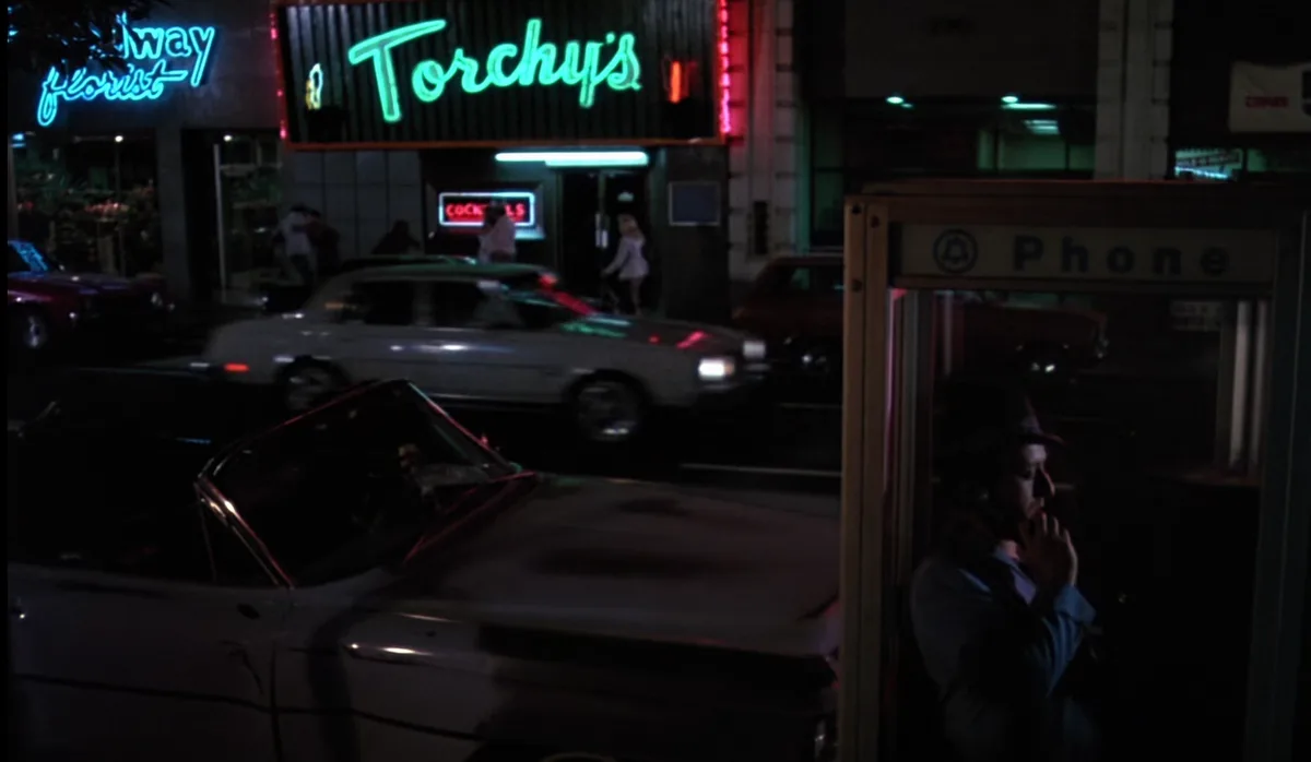 A man in a fedora is on a payphone at night, with neon signs for 'Torchy's' and 'Florist' visible in the background. Cars…