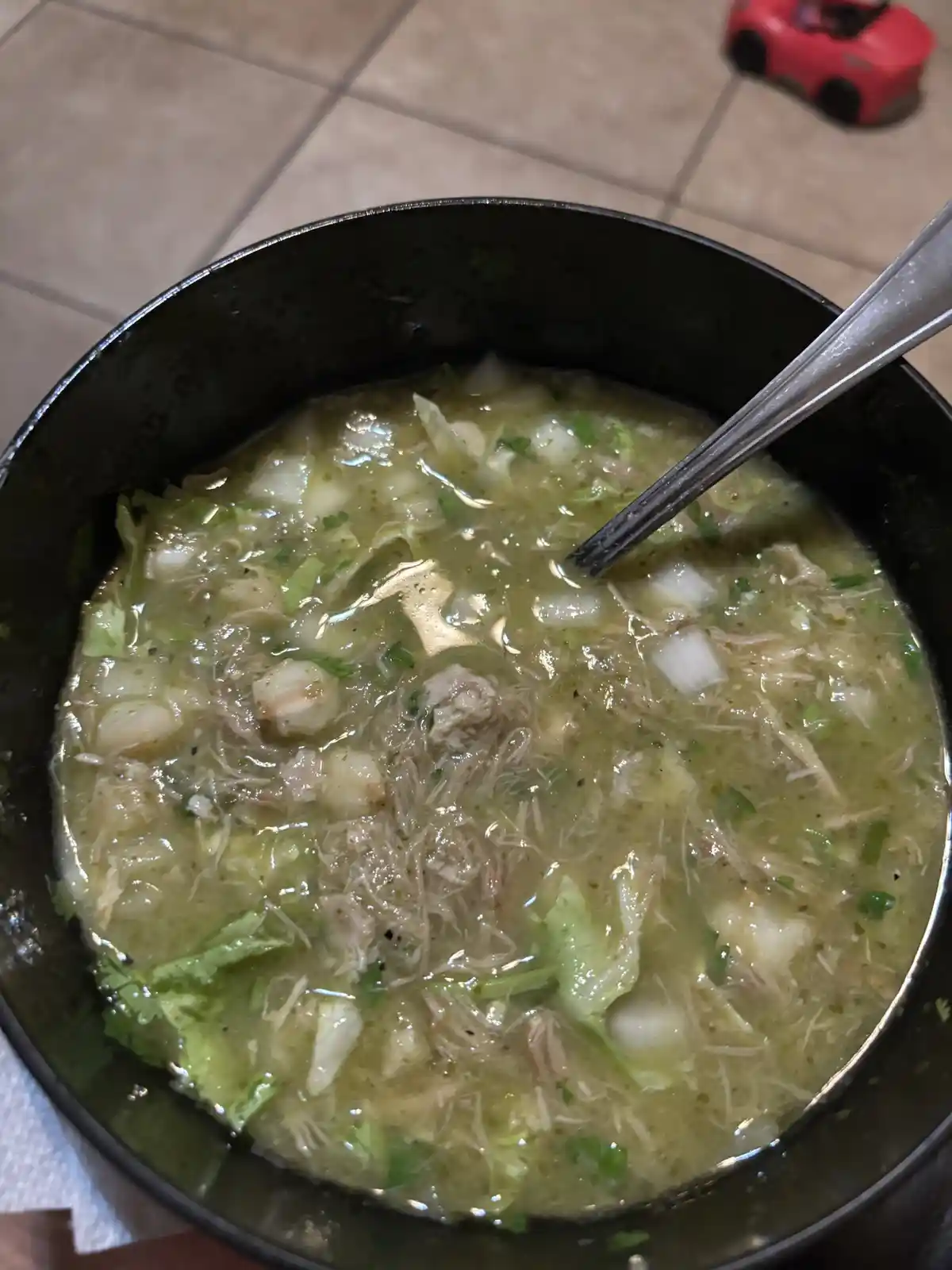 A close-up photo shows a black bowl filled with a greenish, chunky liquid containing shredded meat, cabbage, and herbs, with…