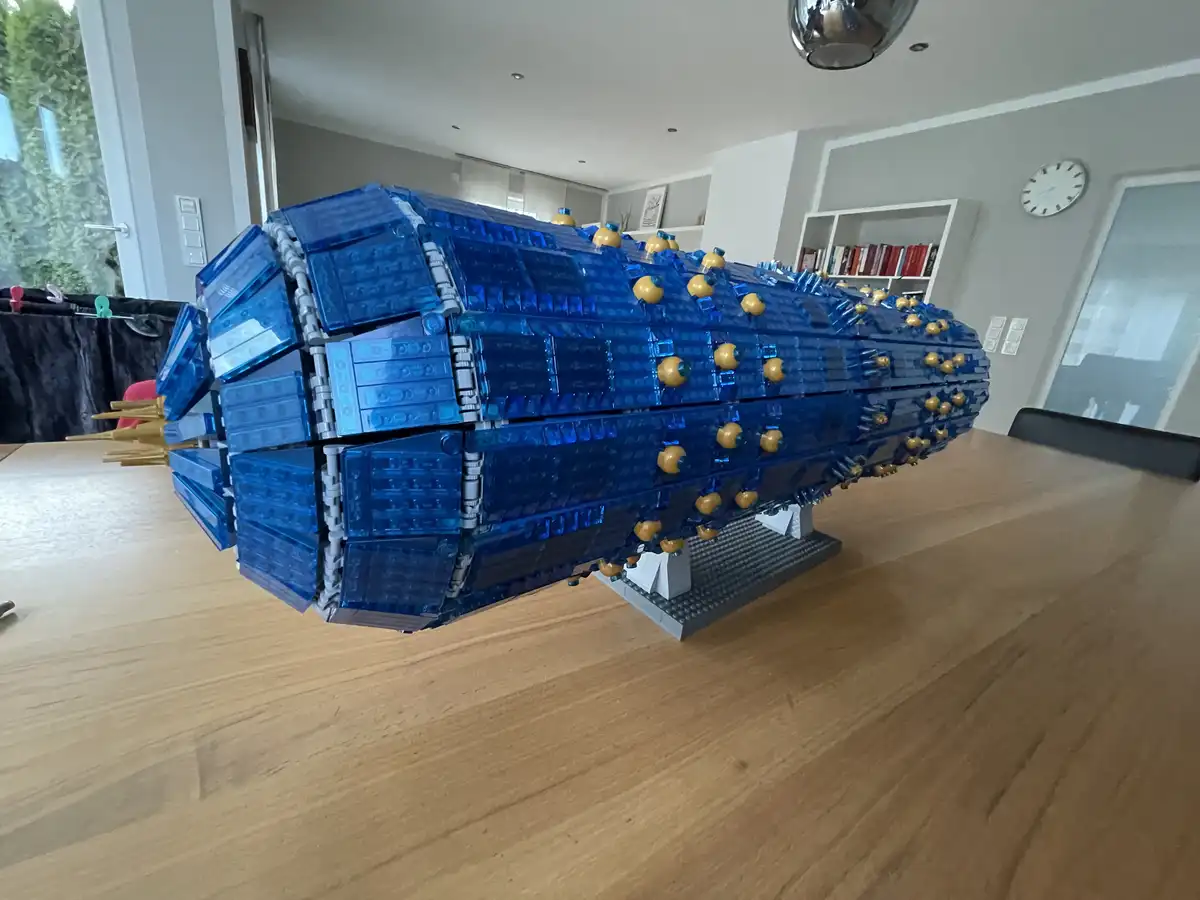 A large, intricate blue Lego spaceship model sits on a wooden dining table in a modern home, featuring gold-colored details…