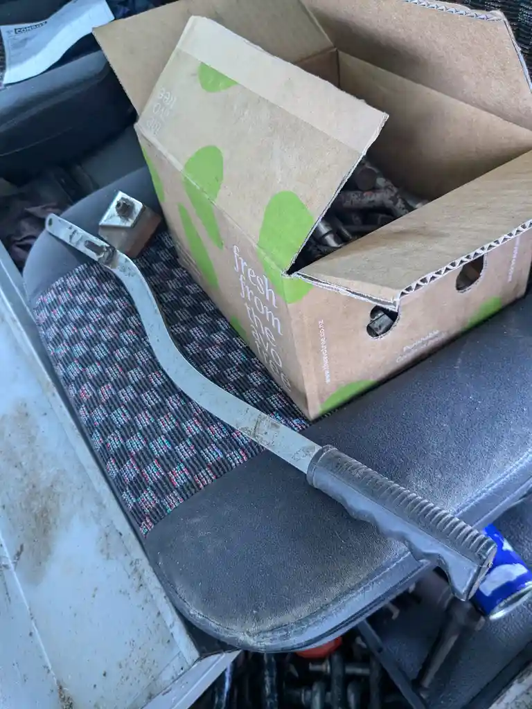A cardboard box labeled 'fresh from the avo tree' sits on a car seat, repurposed to hold rusty metal parts. A large, dirty…