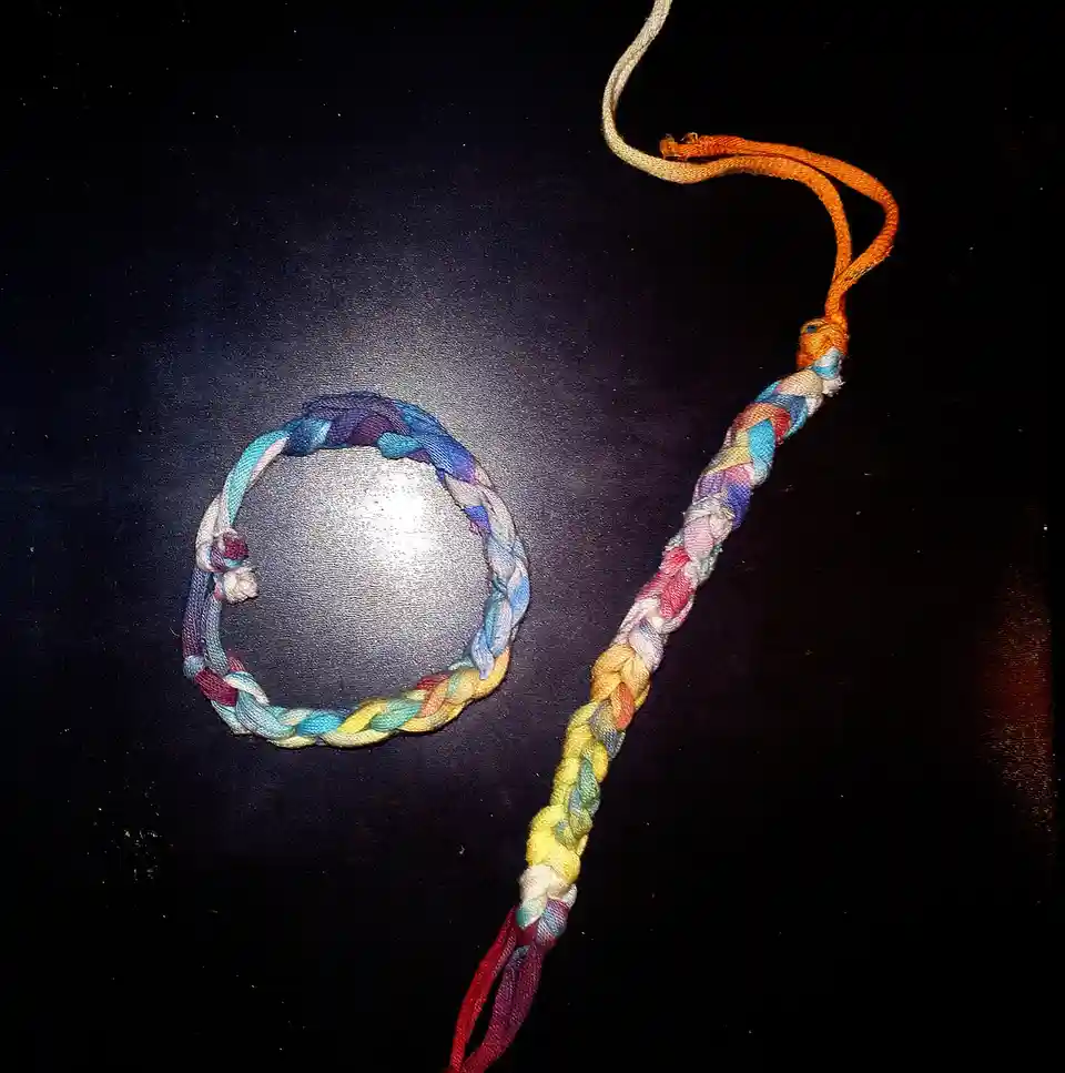 A colorful, hand-braided bracelet and its long, multi-colored cord are laid out on a dark surface, forming the shape of the…