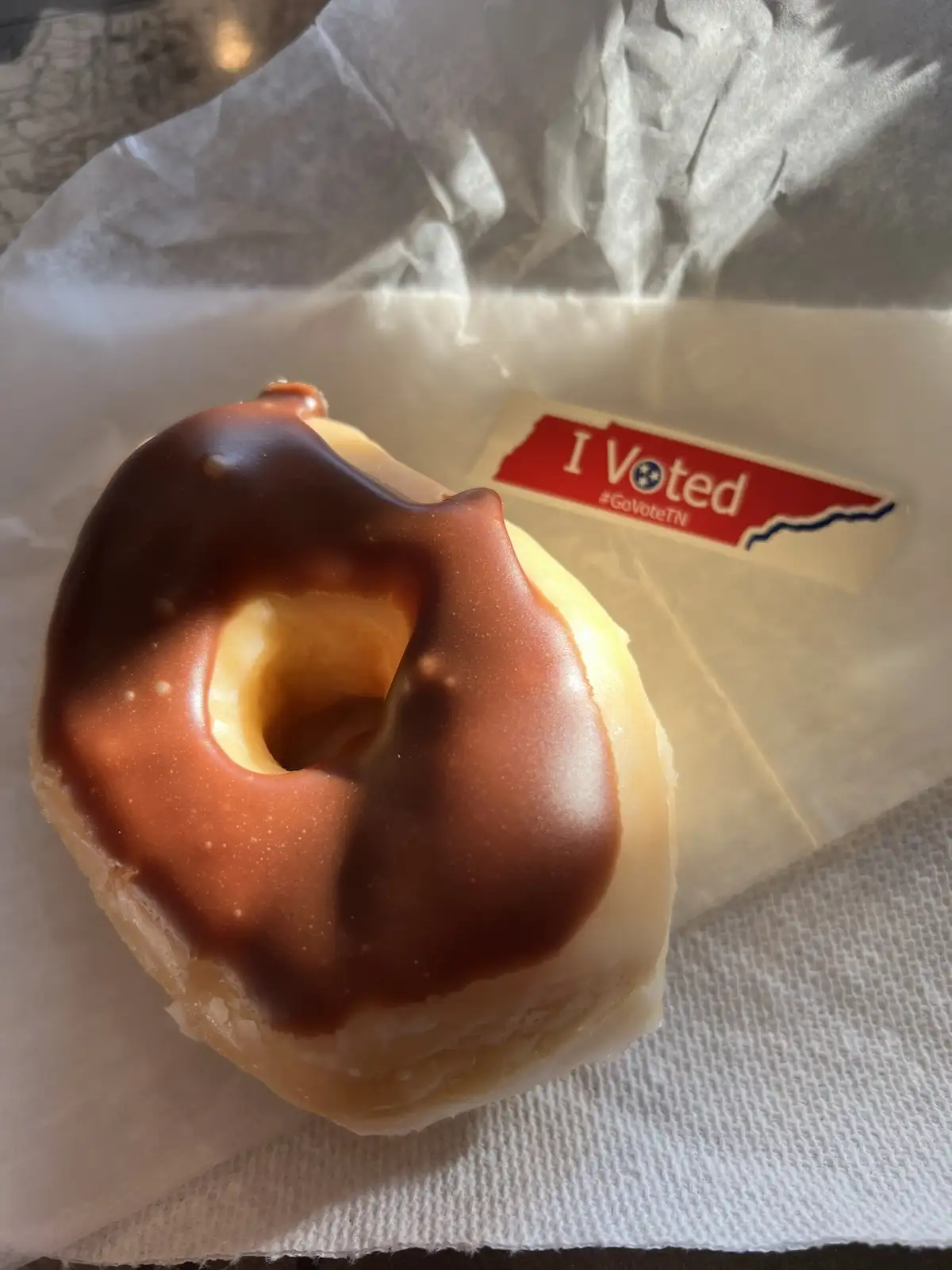 A close-up photo shows a chocolate-glazed donut resting on white paper, with an 'I Voted #GoVoteTN' sticker visible behind…