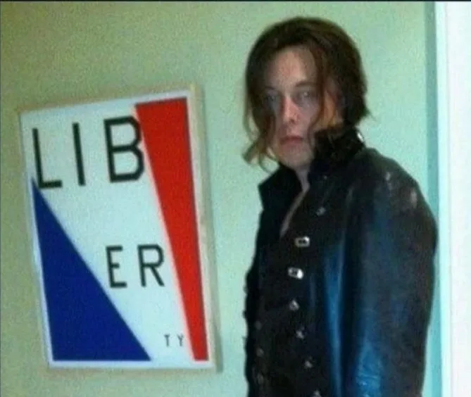 A person with shoulder-length brown hair and glasses, wearing a dark blue leather jacket, stands beside a framed sign. The…