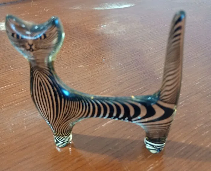A glass figurine of a cat with black and white zebra-like stripes is standing on a wooden surface. The cat has a stylized…