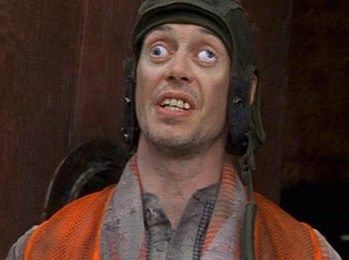 A man wearing a dirty orange vest and a cap with earphones looks upward with wide, startled eyes and an open mouth. The…