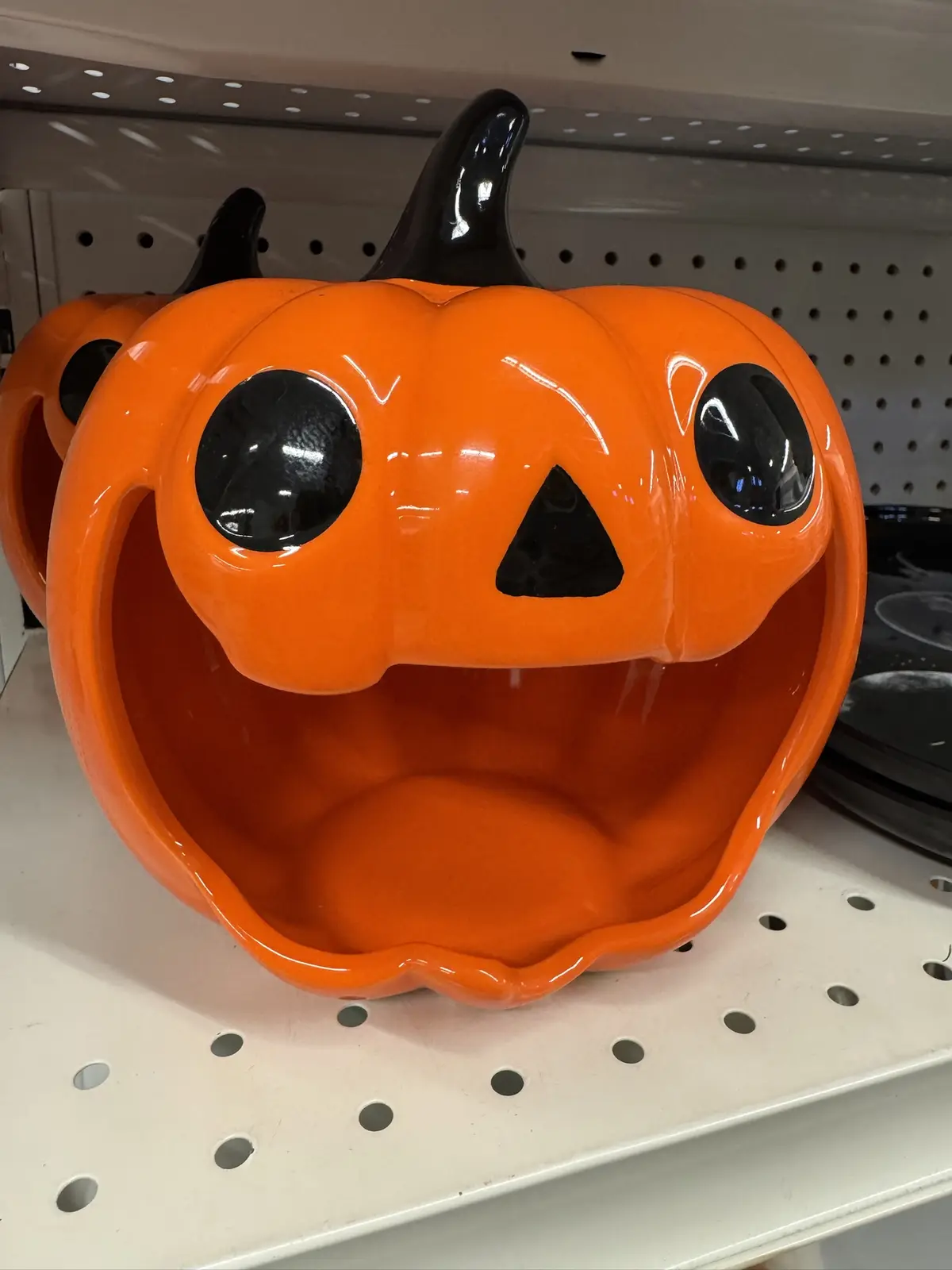 A glossy orange ceramic pumpkin-shaped bowl with large black eyes and a triangular nose sits on a white retail shelf, its…